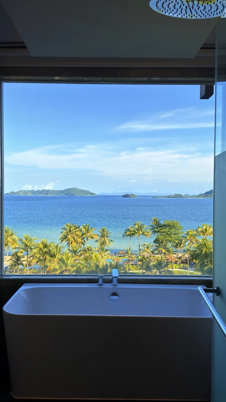 A white bathtub beside a large window overlooking blue water and coconut palms.