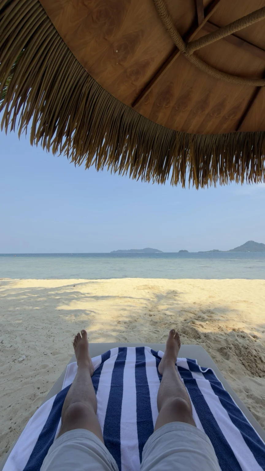 A view from a beach lounger with a blue-and-white striped towel, a thatched umbrella, sand and the sea.