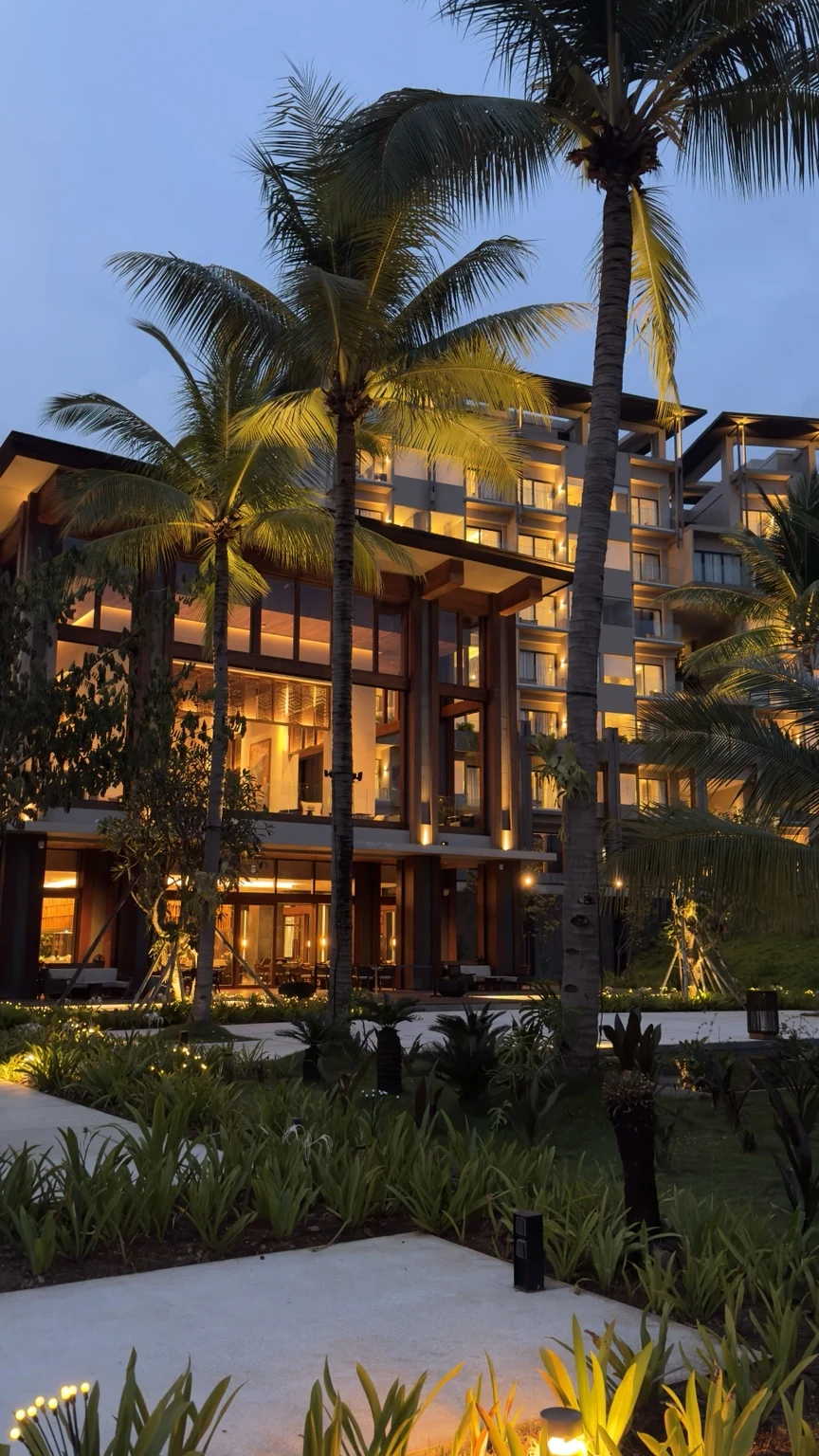 Warmly illuminated resort buildings behind coconut palms and landscaped gardens.