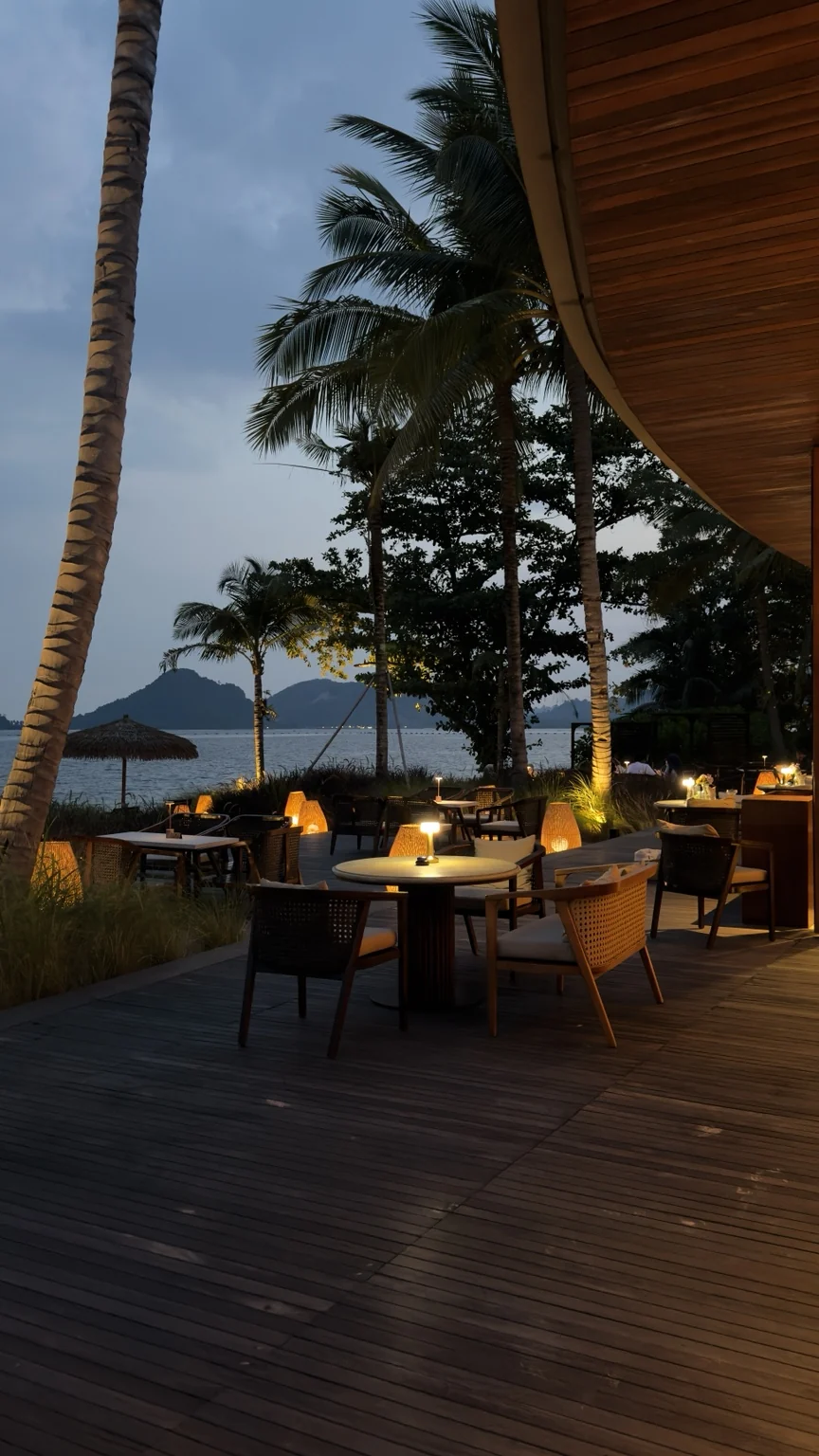 Outdoor dining tables on a deck, lit by small lamps beneath coconut palms at night.