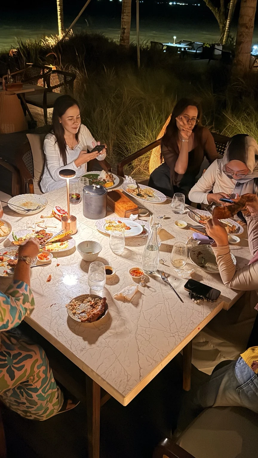Guests sharing an outdoor dinner with several dishes on the table and palms behind them.