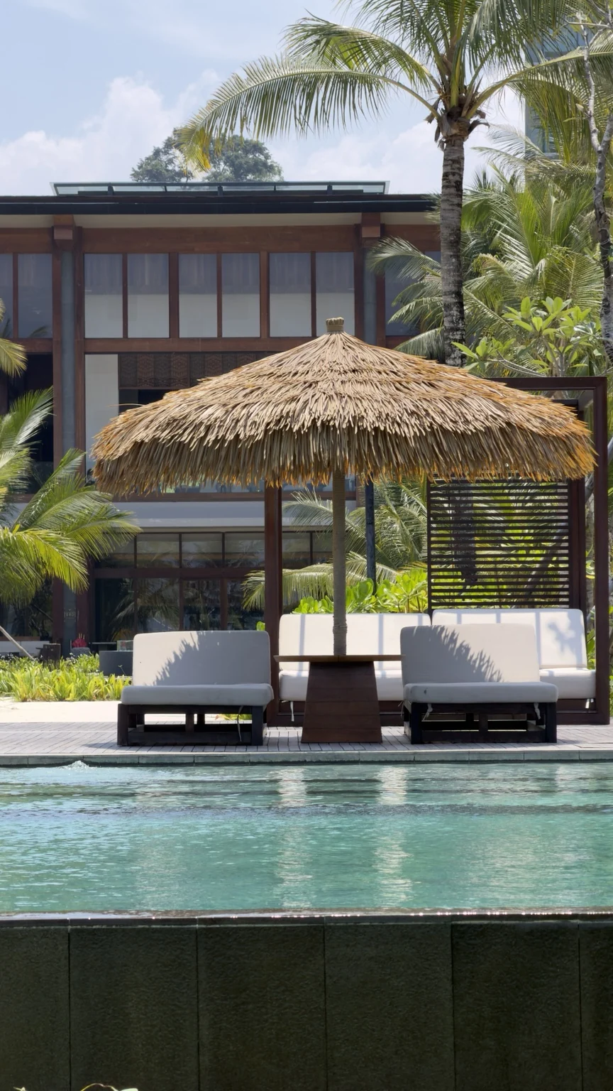 A pool in front of shaded loungers, coconut palms and resort buildings.