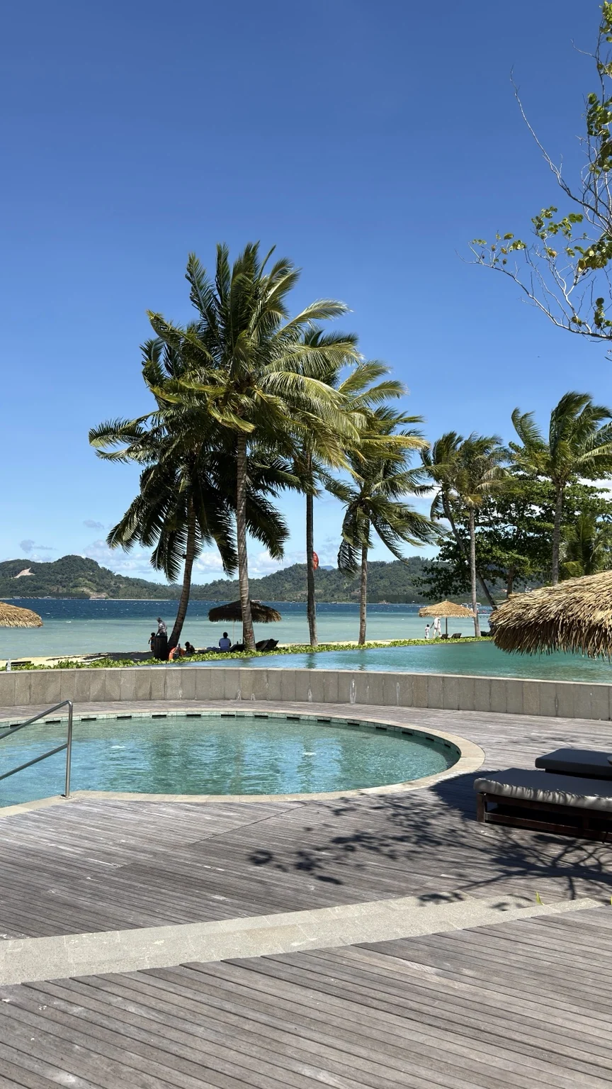 A round pool and wooden deck with loungers, coconut palms and blue water beyond.