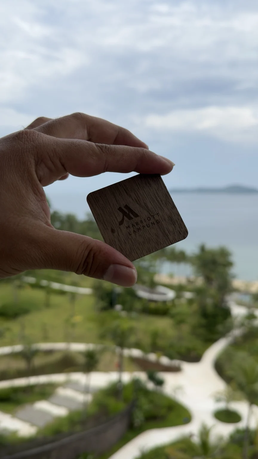 A hand holding a wooden Marriott room key against a backdrop of resort gardens and the sea.