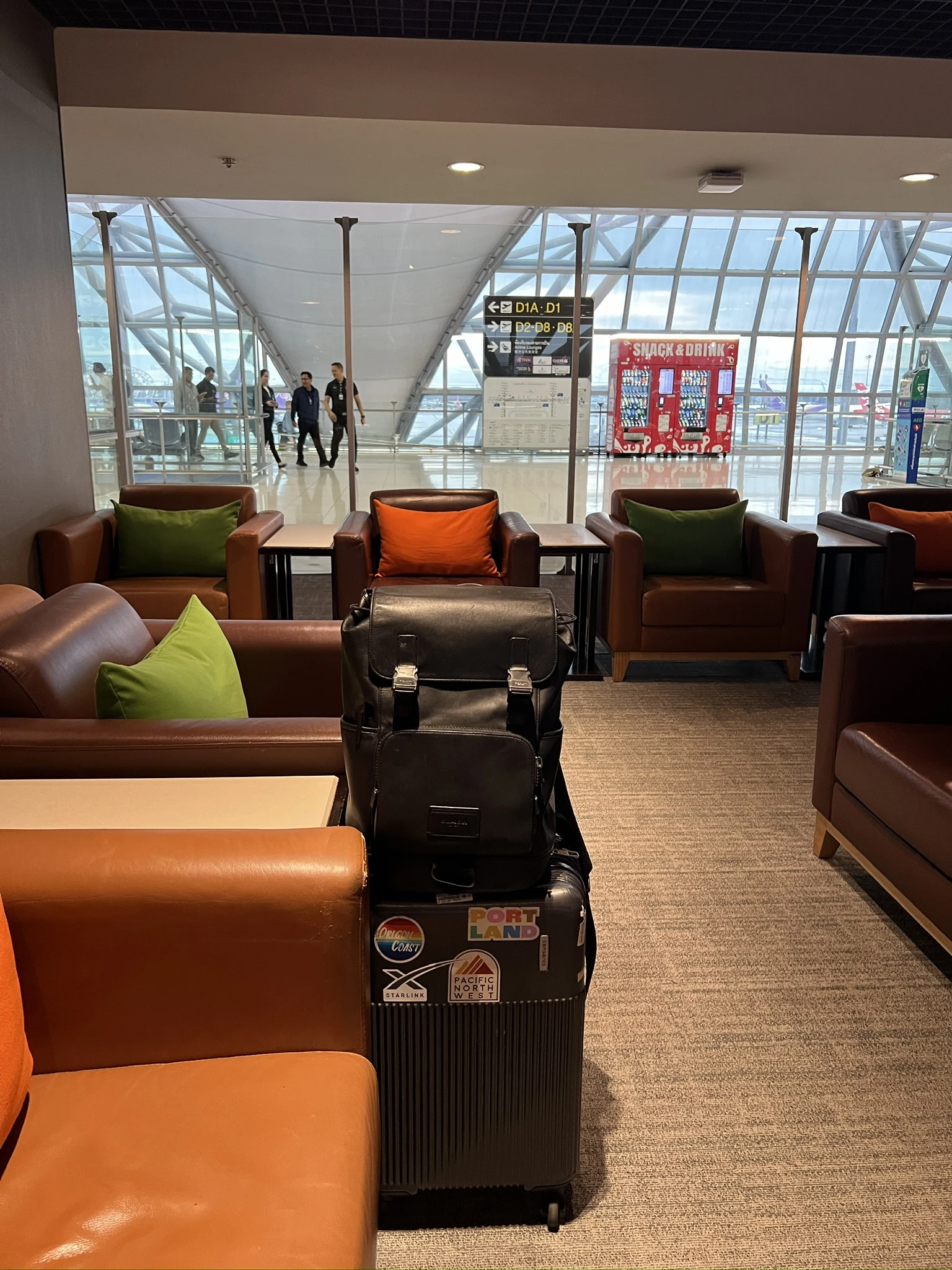 Brown leather lounge chairs with green and orange cushions overlooking the concourse at Bangkok Suvarnabhumi, with my luggage in the foreground.